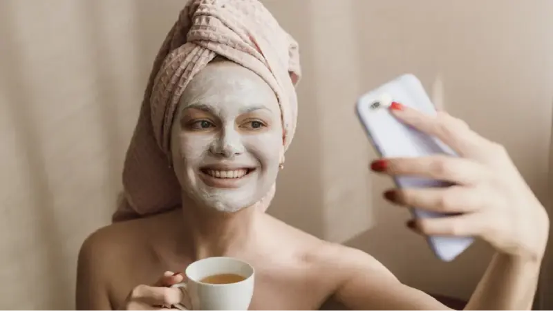 Smiling woman with a face mask and tea, showing how tea rituals aid skin care and relaxation.