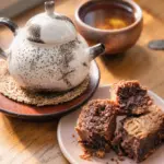 A ceramic teapot, cup of tea and pastries in warm light, representing tea, body awareness and sensory grounding during depression.