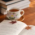 A cup of amber tea beside an open book and autumn leaves on a wooden table.